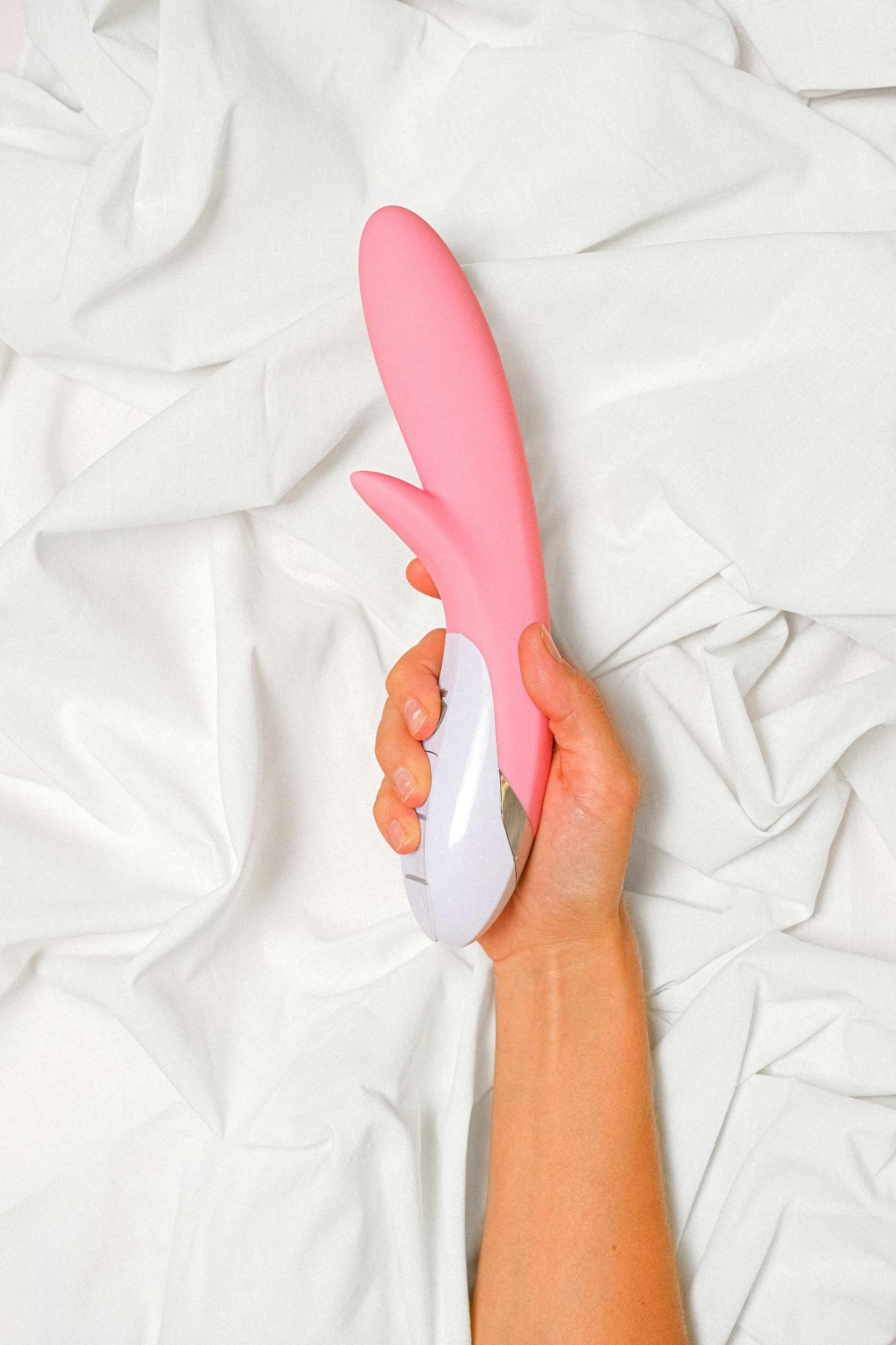 A minimalist shot of a hand holding a pink silicone vibrator on soft, white bed sheets.